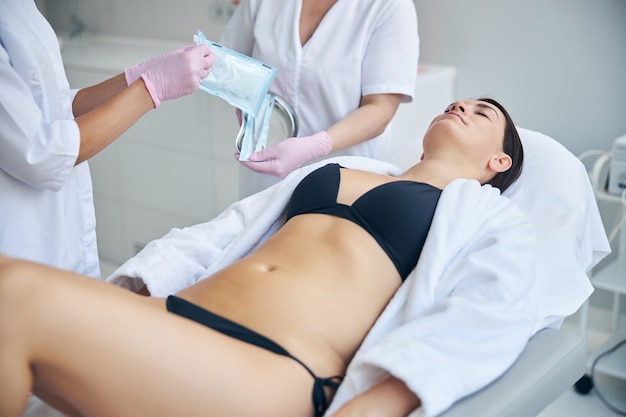 Femme adulte aux cheveux noirs allongée les yeux fermés sur un canapé cosmétique lors d'une procédure médicale