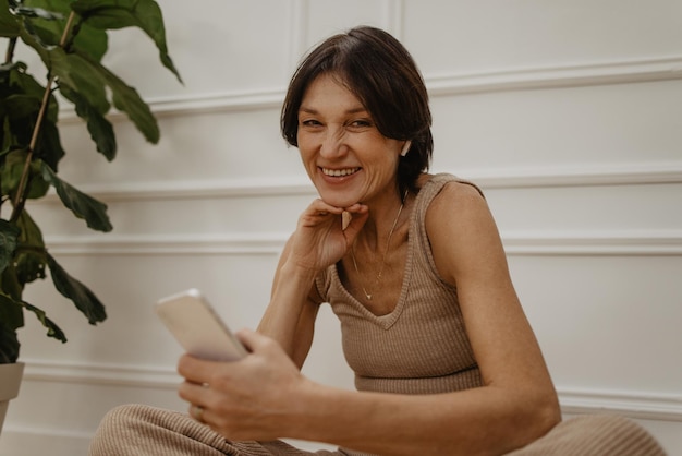 Femme adulte caucasienne passant du temps libre à la maison assise sur le sol dans le salon envoyant des messages texte à l'application mobile sur smartphone