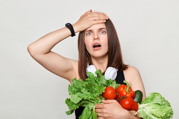 Femme adulte choquée aux cheveux bruns debout isolée sur fond blanc tenant des légumes faisant un geste facepalm regardant la caméra avec de grands yeux