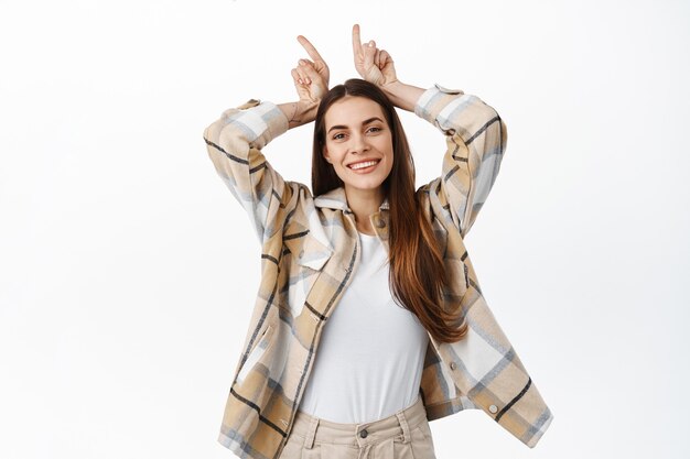 Femme adulte drôle et heureuse jouant, montrant le geste des cornes du diable de taureau et souriant positivement, étant têtue ou déterminée, debout sur un mur blanc