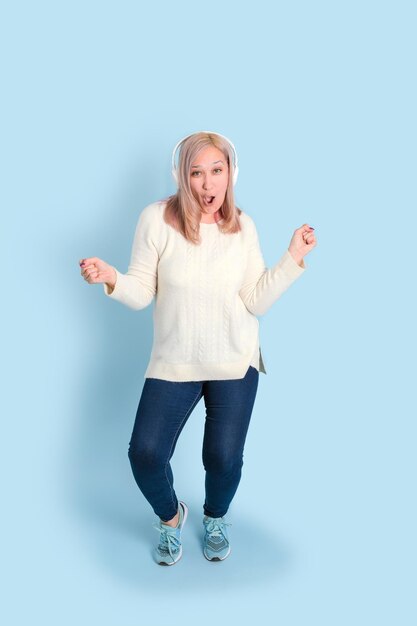 Une femme adulte avec des écouteurs dansant et chantant en écoutant de la musique dans des écouteurs sans fil debout sur un fond bleu. Photo verticale