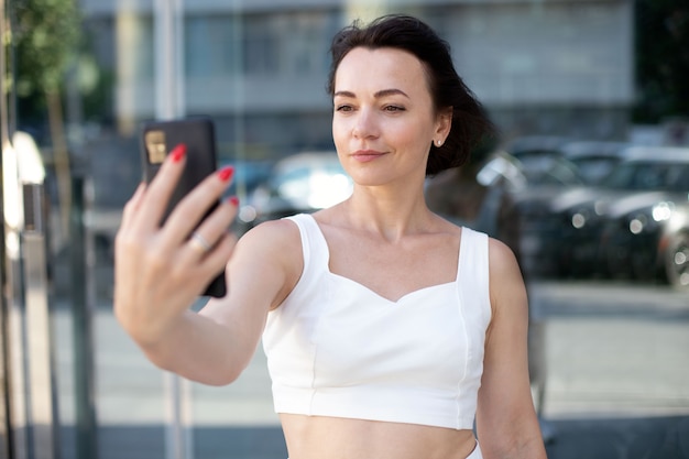 Une femme adulte élégante en blanc prend un selfie au téléphone et sourit en se tenant debout au milieu de la rue urbaine