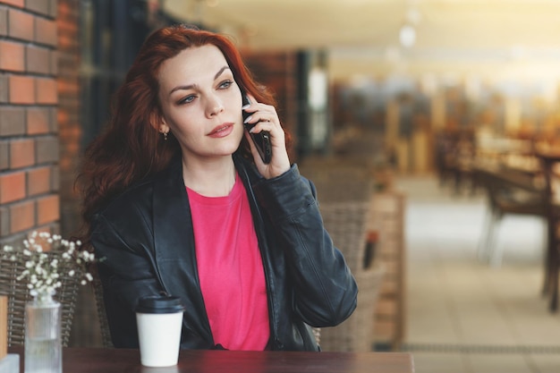 Une femme adulte est assise dans une cafeveranda avec une tasse de café et communique sur un téléphone portable