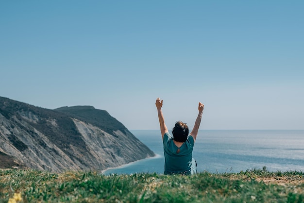 Une femme adulte est assise sur une montagne pieds nus levant les mains vers le ciel et le soleil par une journée ensoleillée sur une montagne sur fond d'océan au printemps vue de dos