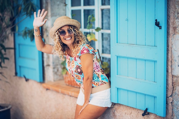 Photo une femme adulte joyeuse dit bonjour à des amis qui sortent d'une maison bleue. touriste heureuse de femmes matures avec des vêtements colorés et un chapeau souriant et profitant d'activités de loisirs en plein air