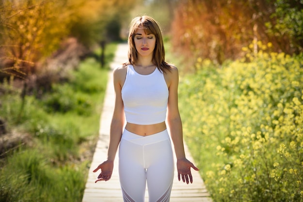 Une femme adulte à mi-âge avec les yeux fermés médite alors qu'elle se tient sur le trottoir au milieu des plantes au parc