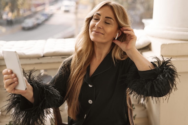 Une femme adulte à la peau claire aime écouter de la musique dans des écouteurs via un smartphone moderne tout en étant assise sur un balcon