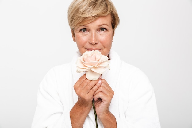 Photo femme adulte à la retraite aux cheveux blonds courts portant une robe de chambre blanche tenant une fleur rose isolée sur un mur blanc