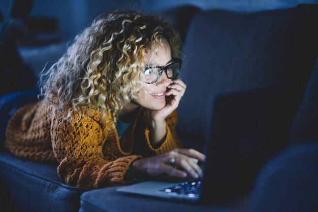 Une femme adulte séduisante sourit et utilise un ordinateur portable et une connexion Internet la nuit, allongée sur le canapé à la maison. Les gens accros à la technologie et aux médias sociaux. Mode de vie féminin heureux