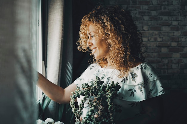 Femme adulte sereine regardant par la fenêtre à la maison seule Vue latérale d'une femme souriante et attentionnée jardin de plantes d'intérieur Heureuse dame solitaire avec des cheveux longs bouclés blonds couleur d'ambiance verte image intérieure