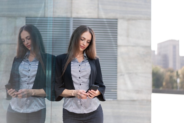 Femme d'affaires de 35 ans vêtue d'une chemise à rayures et d'une veste noire aux cheveux longs debout près d'un smartphone à usage extérieur d'un immeuble de bureaux