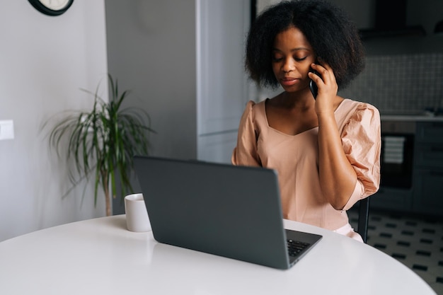 Femme d'affaires africaine bouclée dans des vêtements décontractés travaillant à partir d'un bureau à domicile distant faisant un appel téléphonique à un client potentiel ayant une conversation assis au bureau