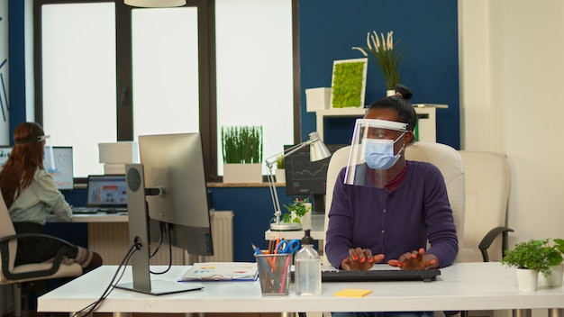 Femme d'affaires africaine rédigeant des rapports sur ordinateur dans un bureau de société financière très fréquenté travaillant avec des collègues respectant la distance sociale et portant une visière et un masque de protection.