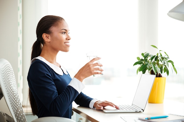 Femme d'affaires africaine en robe assise sur un fauteuil avec de l'eau près de la fenêtre au bureau