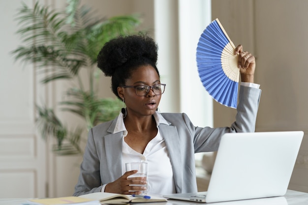 Une femme d'affaires africaine surchauffée boit de l'eau d'un ventilateur assis au bureau avec un ordinateur portable ou à la maison