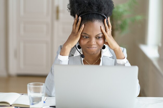 Femme d'affaires afro agacée en colère crier, porter des écouteurs parler à un mauvais client de travailler sur un ordinateur portable