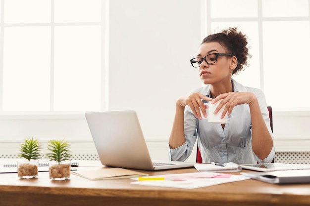 Femme d'affaires afro-américaine détendue réfléchie assise sur le lieu de travail dans un bureau moderne avec une tasse de café