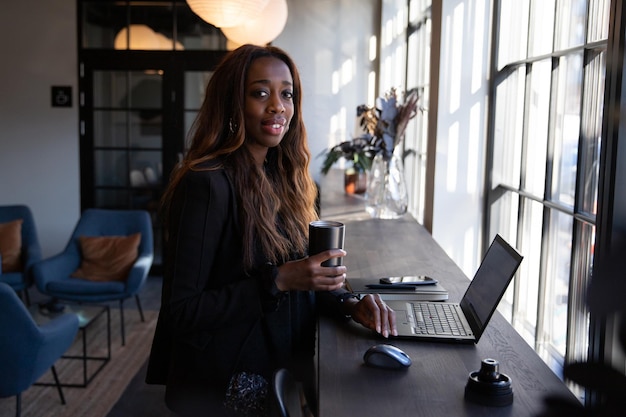 Femme d'affaires afro-américaine souriante buvant du café au bureau et travaillant avec un ordinateur portable