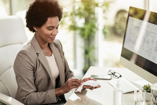 Femme d'affaires afro-américaine utilisant un gel antiseptique pour désinfecter sa souris d'ordinateur alors qu'elle était assise seule dans un bureau au travail.