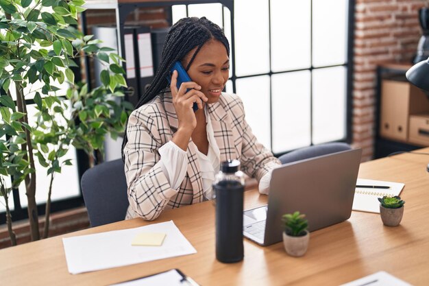 Femme d'affaires afro-américaine utilisant un ordinateur portable parlant sur un smartphone au bureau