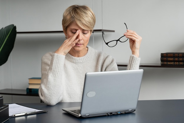 Une femme d'affaires d'âge moyen assise à son bureau enlève ses lunettes à cause de la douleur dans ses yeux et