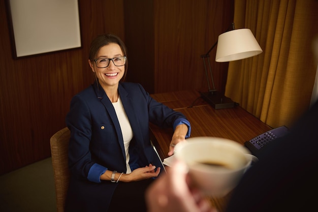 Femme d'affaires d'âge moyen, mignonne souriante, sourire à pleines dents, regardant son collègue, son partenaire autour d'une tasse de café, se relaxant dans une chambre d'hôtel après de longues négociations au bureau lors d'un voyage d'affaires