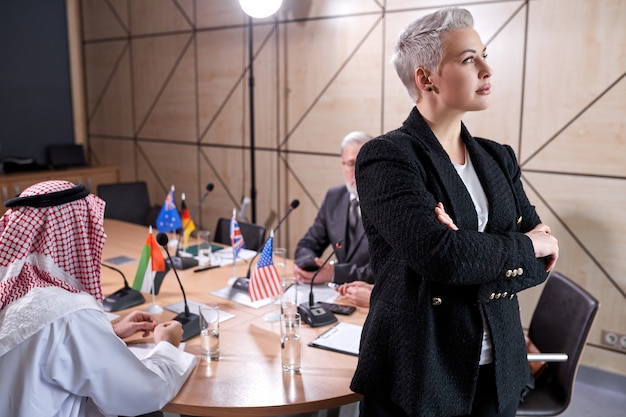 Femme d'affaires âgée avec des cheveux courts élégants en tenue de soirée posant dans la salle de conférence lors d'une réunion avec un groupe international de politiciens assis au bureau en arrière-plan, elle regarde sur le côté. portrait