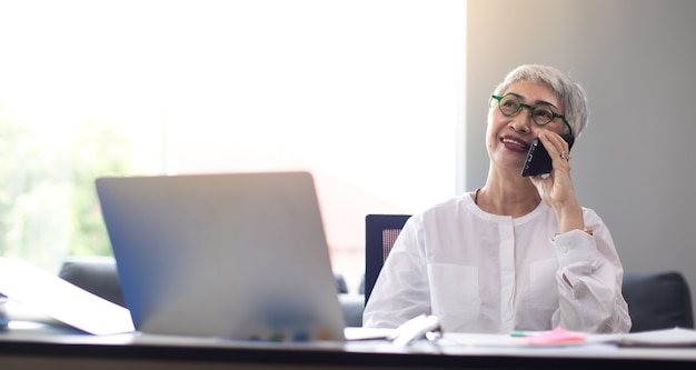 Femme d'affaires âgée séduisante travaillant sur un ordinateur portable et passant un appel téléphonique au bureau à domicile.