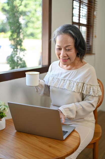 Femme d'affaires asiatique âgée prospère travaillant à domicile en sirotant un café tout en utilisant un ordinateur portable