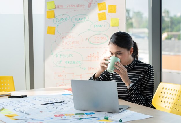 Femme d'affaires asiatique boire du café et de la vidéo en ligne avec ordinateur portable et tableau de bord au bureau