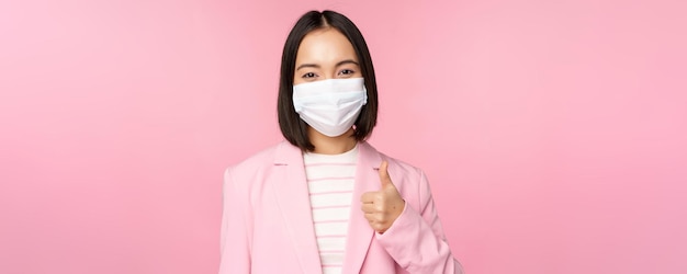 Femme d'affaires asiatique en costume et masque médical montrant les pouces vers le haut recommandant de porter un équipement de protection individuelle au bureau pendant la pandémie de covid19 fond rose