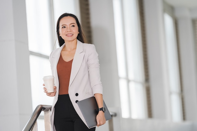 Une femme d'affaires asiatique dans un costume décontracté tient une tasse de café à emporter dans le bâtiment de l'entreprise