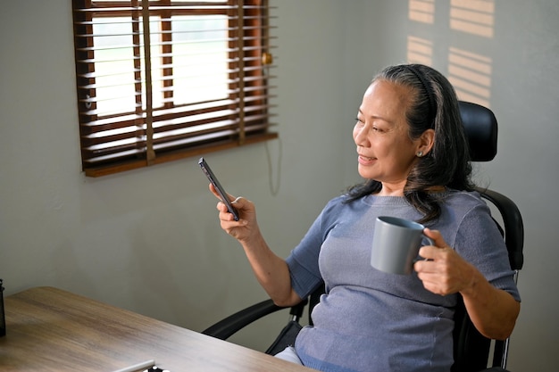 Femme d'affaires asiatique détendue en sirotant un café et en utilisant son téléphone à son bureau