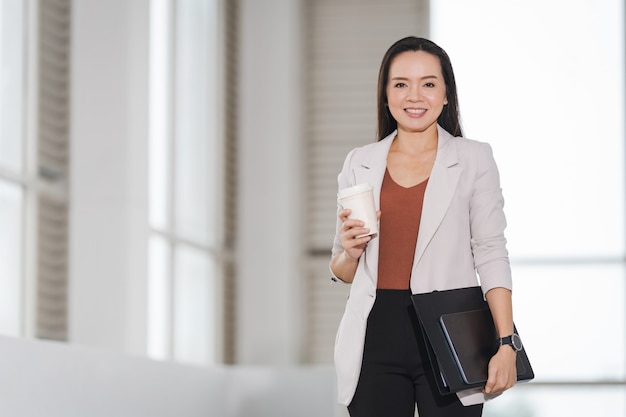 Une femme d'affaires asiatique heureuse et prospère tient une tasse de café à emporter et un document dans la construction d'entreprises