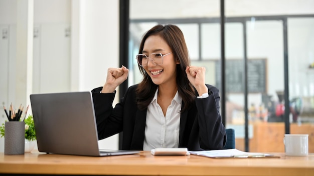 Femme d'affaires asiatique joyeuse montrant des mains triomphantes souriant et regardant l'écran d'un ordinateur portable