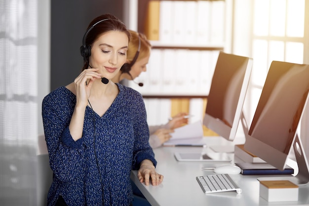 Femme d'affaires asiatique ou latino-américaine parlant par casque dans un bureau ensoleillé. Centre d'appels et groupe de personnes diverses dans les affaires.