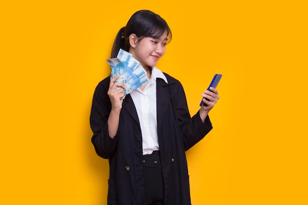 Femme d'affaires asiatique montrant de l'argent et tenant un téléphone portable isolé sur fond jaune