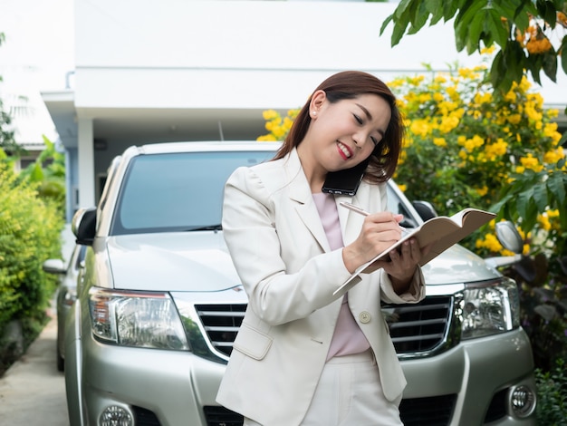 Femme d'affaires asiatique parlant sur téléphone mobile et prendre des notes contre une voiture.