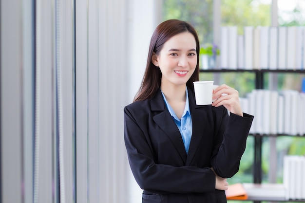 Une femme d'affaires asiatique qui porte une chemise bleue et un costume noir tient une tasse de café à la main et sourit