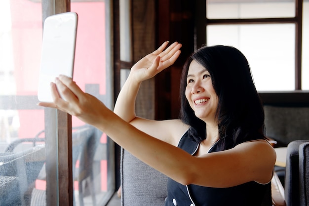 Une femme d'affaires asiatique souriante passe des appels vidéo sur les réseaux sociaux avec une tablette dans un café. Elle parle pour le travail et la technologie dans l'espace de travail au bureau ou à domicile pendant l'épidémie de COVID-19