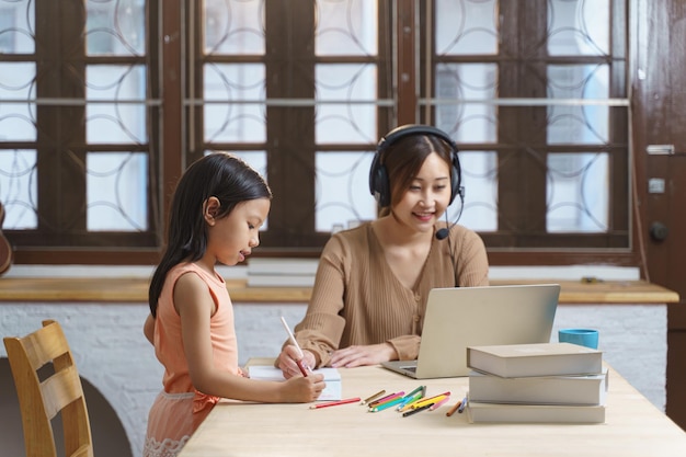 Photo une femme d'affaires asiatique travaillant sur un ordinateur portable assis au bureau à domicile pendant que sa fille en tant qu'étudiante fait ses devoirs
