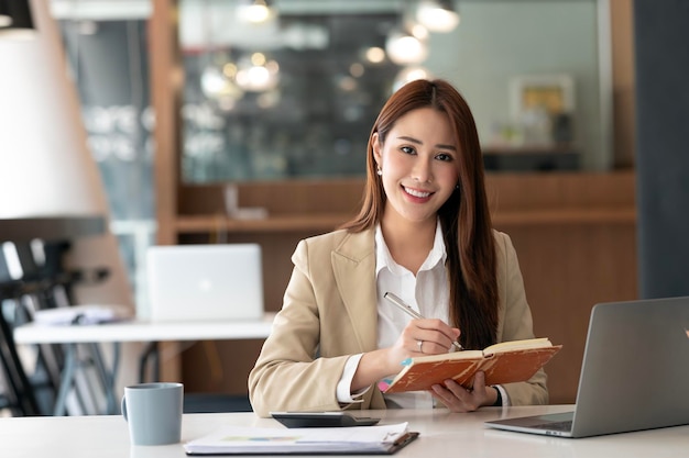 Une femme d'affaires asiatique travaille avec son calculateur portable et des documents dans le bureau en souriant à la caméra