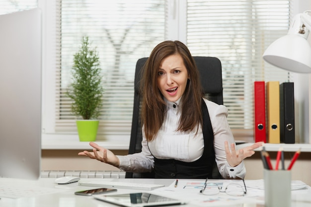 Une femme d'affaires assez en colère en costume assise au bureau avec une tablette, travaillant sur un ordinateur avec un moniteur moderne avec des documents dans un bureau léger, jurant et criant, résolvant des problèmes, concept d'émotion.