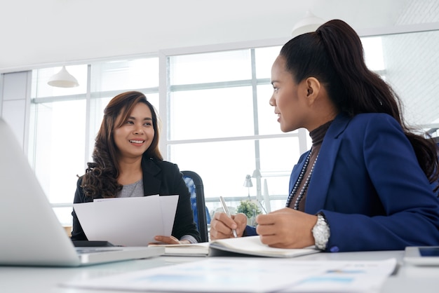 Femme d'affaires assez souriante discutant d'idées commerciales avec un collègue et prenant des notes dans le planificateur