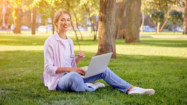 Une femme d'affaires assise sur l'herbe dans un parc d'été en utilisant un ordinateur portable. Une personne d'affaires travaillant à distance en plein air.