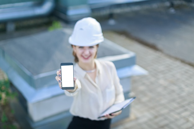 Femme d'affaires attrayante en blouse blanche, montre, casque et jupe noire se tient sur le toit et montre un smartphone vide