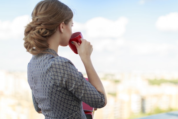 Femme d'affaires attrayante en robe à motifs se tenir sur le toit et tenir le dossier papier et la tasse rouge