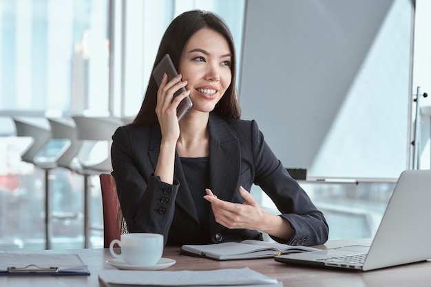 Femme d'affaires au bureau seule assise à table avec une tasse de café chaud parlant sur smartphone avec un collègue souriant joyeux