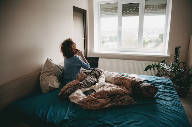 Femme d'affaires aux cheveux bouclés, parler au téléphone dans la chambre tout en tenant un ordinateur portable