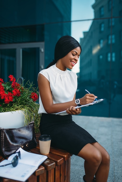 Femme d'affaires avec bloc-notes assis sur le banc pendant la pause en face de l'immeuble de bureaux